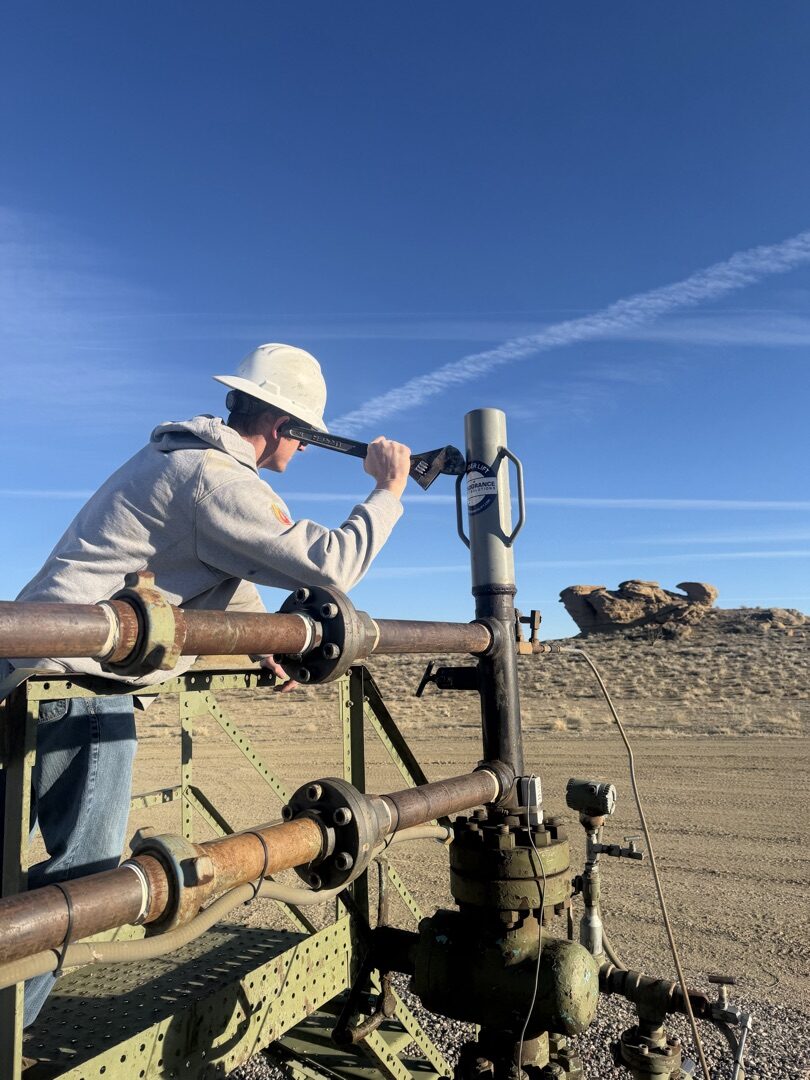 Operator using a crescent wrench pressed against a wellhead pipe to listen for plunger activity — the old way